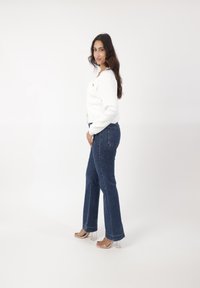 Witte geribde top met lange mouwen en een knoop aan de voorkant, gecombineerd met donkerblauwe wijde jeans en doorzichtige sandalen met hakken, zijwaarts gepositioneerd.