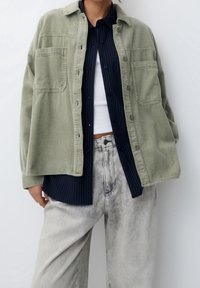 Chaqueta de camisa de pana verde oliva con dos bolsillos frontales, superpuesta a una camisa de rayas azul marino. Combinada con jeans desgastados de color gris claro.
