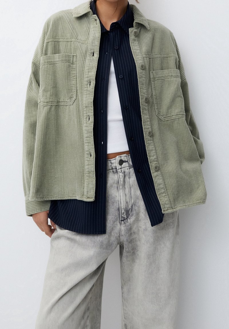 Chaqueta de camisa de pana verde oliva con dos bolsillos frontales, superpuesta a una camisa de rayas azul marino. Combinada con jeans desgastados de color gris claro.