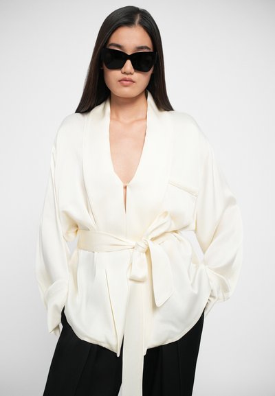 Blusa in satin bianco con scollo a V profondo, maniche larghe e vita legata. Abbinata a pantaloni neri e occhiali da sole neri oversize.