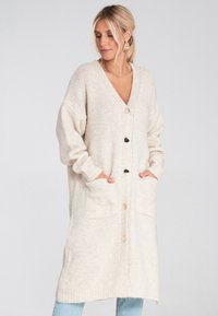 Kobieta z blond włosami związanymi w niski kucyk, nosząca złote kolczyki i długi beżowy kardigan zapinany na guziki z kieszeniami, ręce włożone w kieszenie.