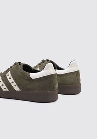 Zapatillas de ante verde con acentos blancos, suelas marrones y tres ranuras con tachuelas doradas a lo largo de los lados. Diseño de perfil bajo.