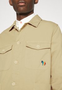 Chaqueta beige con botones, fabricada en algodón, que presenta dos bolsillos en el pecho y un pequeño bordado multicolor de un caballo en el bolsillo izquierdo.