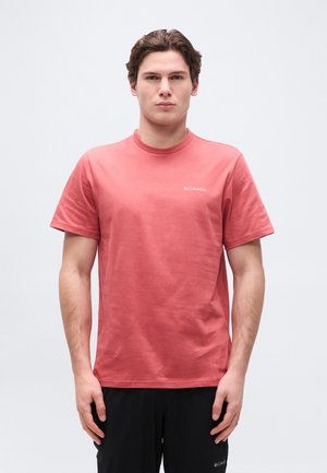 Fiatal férfi korallszínű Columbia pólóban és fekete nadrágban, egyenesen áll egy egyszínű, világos háttér előtt.