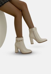 Vita ankelbooties gjorda av slät läder med klack, med dragkedjestängning och ett dekorativt band med guldaccent.