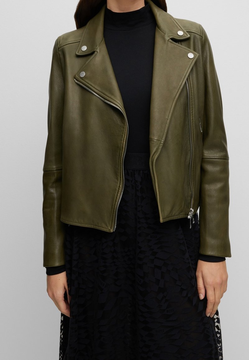 Femme portant une veste de motard en cuir vert olive sur un haut noir à col montant et une jupe noire texturée, debout devant un fond uni.