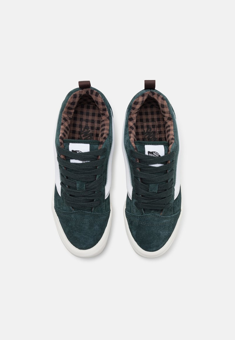 Baskets en suède vert avec des rayures blanches sur les côtés et des lacets vert foncé. L'intérieur présente un motif à carreaux marron et noir. Semelles en caoutchouc solides.