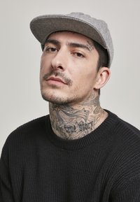 Grå følt caps, svart strikkegenser, markert hals tatovering med skrift, nærbilde av ansiktet, nøytral bakgrunn.