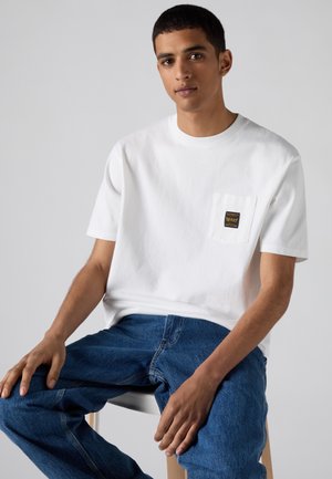 Junger Mann mit kurzen Haaren, der ein weißes Kurzarmhemd mit Taschenlogo und blaue Jeans trägt und auf einem Hocker vor schlichtem Hintergrund sitzt.