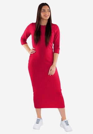 Kobieta o długich ciemnych włosach, ubrana w dopasowaną czerwoną sukienkę midi z rękawami do połowy przedramienia i białe trampki, stoi z jedną ręką na biodrze.