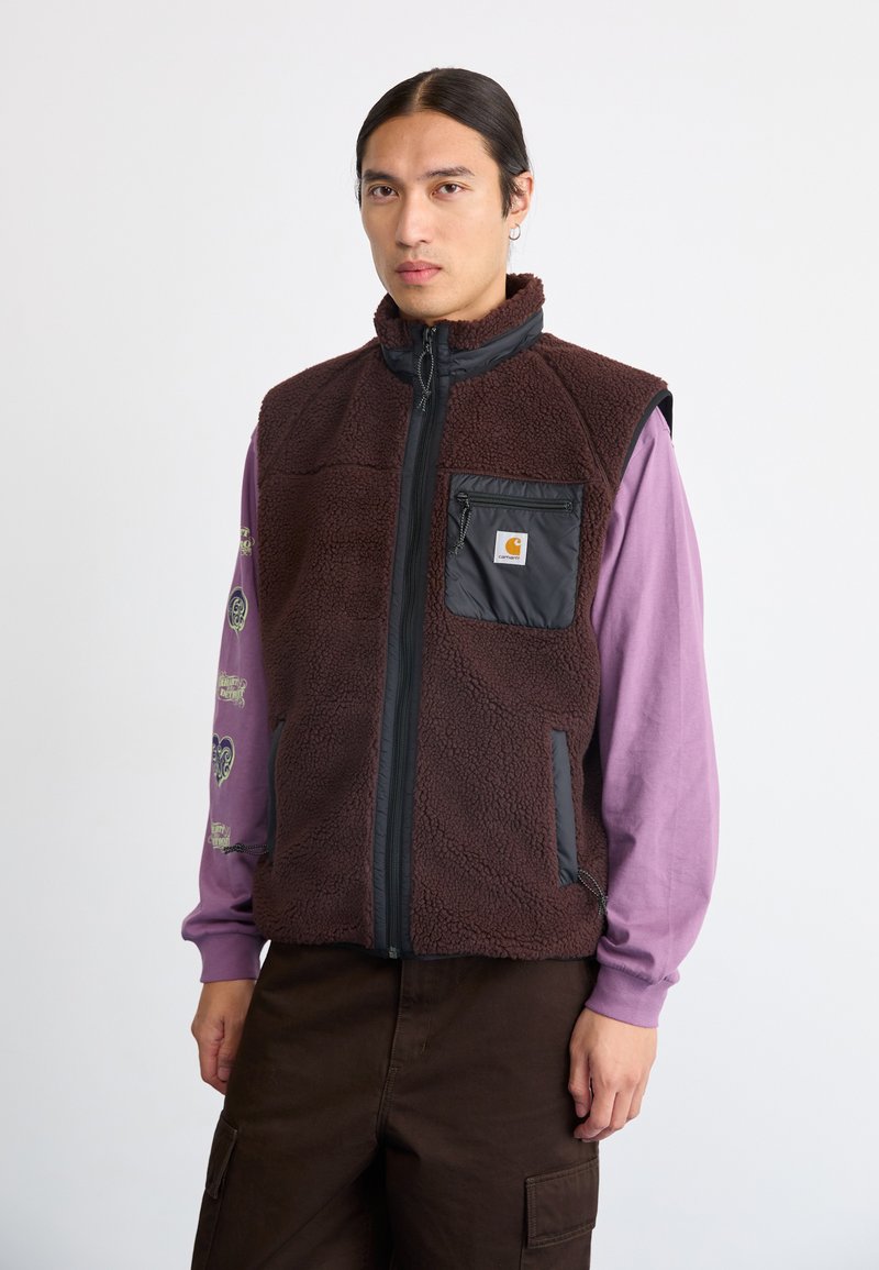Karma brūna fleeces veste ar rāvējslēdzēja priekšpusi, augstu apkakli un melnu neilona kabatu. Nēsāta virs garām piedurknēm violetas krāsas krekla ar rakstainām piedurknēm.