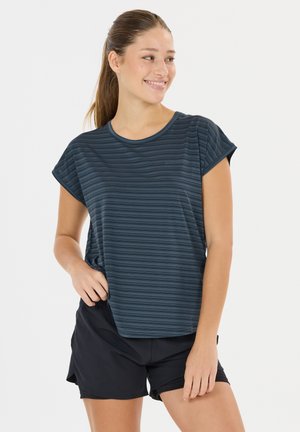 Nuori nainen hymyilee, seisoo käsi taskussa, yllään tummansininen raidallinen T-paita ja mustat urheilushortsit valkoisella taustalla.