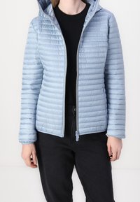 Lyseblå, quiltet pufferjakke med hætte og lynlås foran. Har en struktureret overflade og elastiske manchetter. Båret over en sort top.
