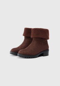 Cizme scurte din suede maro, cu un manschet din blană sintetică, toc pătrat și talpă din cauciuc cu un model de profil pentru aderență.