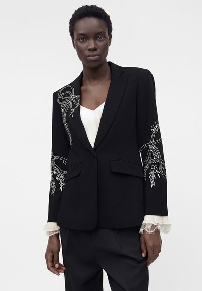 Μαύρο κοσμημένο σακάκι με λευκή κεντητή λουλούδια και φιόγκο στους ώμους και τα μανίκια, φορεμένο πάνω από λευκό πουκάμισο με δαντελωτά μανίκια.