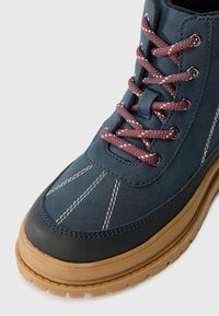 Stivaletto alla caviglia blu in suede con lacci intrecciati rossi e grigi, cuciture bianche, suola in gomma e punta rotonda. Finitura testurizzata sulla parte superiore.