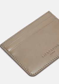 Skórzany portfel na karty w jasnobeżowym kolorze z czterema przegródkami na karty, wytłoczonym logo i starannym przeszyciem na krawędziach. Gładka faktura i minimalistyczny design.