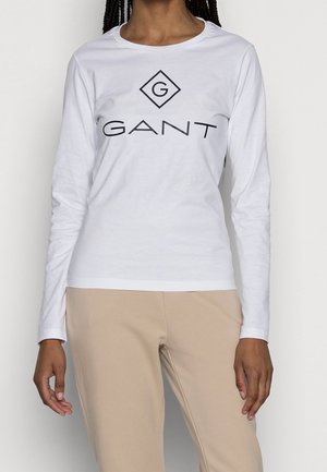 Personne portant une chemise blanche à manches longues avec un logo "GANT" et un pantalon beige, montrée du cou aux hanches.