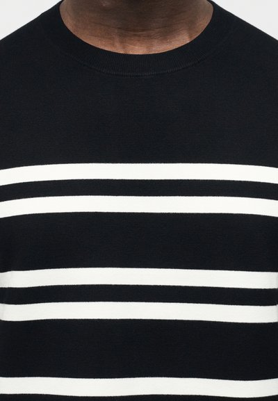 Εγγύς πλάνο ενός ατόμου που φορά μαύρο πουλόβερ με τρεις λευκές οριζόντιες ρίγες στο στήθος.