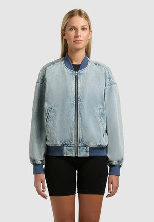 Fiatal nő áll, világoskék farmer bomberdzsekit visel sötétkék mandzsettákkal és gallérral, fekete rövidnadrággal egy egyszínű háttér előtt.