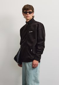 Ung mand iført sort lynlåstrøje, lyseblå jeans, sorte solbriller og holder en sort taske mod en ensfarvet baggrund.