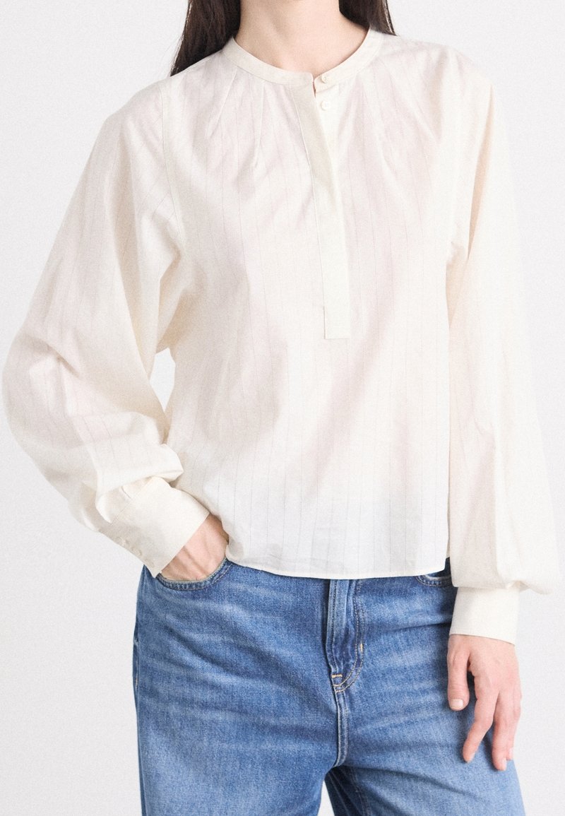 Femme portant un chemisier ample de couleur crème avec des manches bouffantes et un jean bleu, une main dans la poche, sur un fond uni.