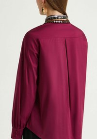 Long-sleeved maroon blouse featuring a textured collar with shiny accents, back pleat detail, and buttoned cuffs. Smooth fabric texture.