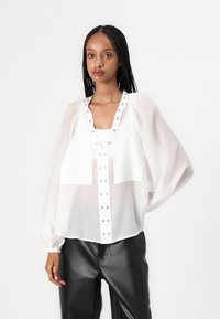 Blusa bianca traslucida con maniche lunghe a sbuffo, chiusura frontale con bottoni decorata con borchie argentate, abbinata a pantaloni di pelle nera.