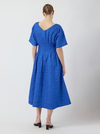 Blaues, strukturiertes Kleid mit V-Ausschnitt, kurzen Ärmeln und gerafftem Rücken. Das Design verfügt über einen fließenden Rock, der bis zur Mitte der Wade reicht.