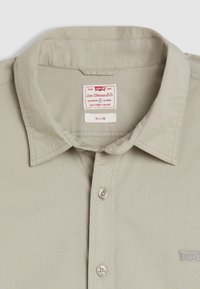 Chemise slim-fit beige avec un col pointu, fermeture à boutons sur le devant et un petit logo brodé. Comprend une étiquette d'entretien à l'intérieur du col.