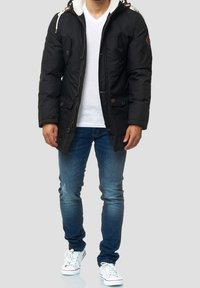 Parka noire isolée avec une capuche bordée de fausse fourrure, détails à boutons, et deux poches avant, portée avec un jean bleu et des baskets blanches.