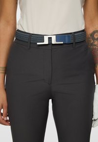 Persona che indossa pantaloni scuri e una camicia bianca con una cintura tessuta blu scuro dotata di una fibbia rettangolare metallica, con un braccio tatuato visibile.