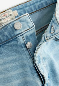 Jean en denim bleu clair à coupe slim. Comprend des boutons en métal argenté et des coutures contrastantes orange sur une texture de tissu lisse.