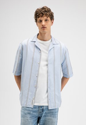 Mann mit kurzen, lockigen Haaren trägt ein hellblaues, kurzärmeliges, gestreiftes Hemd über einem weißen T-Shirt und hellblaue Jeans, steht vor einem schlichten Hintergrund.