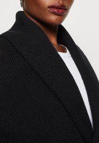 Cardigan nero in maglia con ampio colletto e tessuto testurizzato, con uno strato interno bianco liscio visibile al collo.