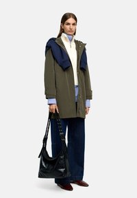 Olivew Green parka z zamkiem błyskawicznym, nałożona na kremowy sweter i niebieską koszulę. Czarna skórzana torebka na ramię, szerokie dżinsowe spodnie, brązowe buty.