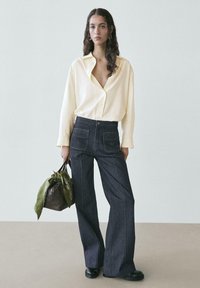 Weit geschnittene, dunkle Jeanshosen mit Kontrastnähten, kombiniert mit einem gelben Hemd zum Knöpfen. Trägt eine braune Tasche mit grünem Schalsakzent.