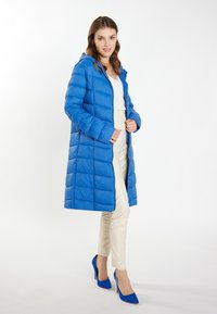 Palton puffer lung, albastru, cu glugă, cu un design matlasat, fermoar frontal și buzunare laterale. Asortat cu pantaloni de culoare crem și tocuri albastre.