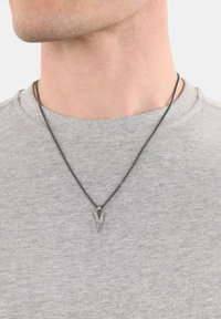 Czarny łańcuszek na szyi z trójkątnym metalowym wisiorkiem z geometrycznymi detalami, noszony na szarym t-shircie z okrągłym dekoltem.