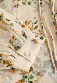 Tissu beige transparent avec des points texturés, imprimé floral, poignet froncé avec un bouton en nacre et des cordons au cou à nouer.