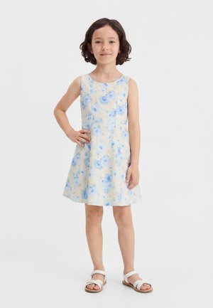 Fille aux cheveux courts et bruns portant une robe blanche sans manches avec des fleurs bleues et des sandales blanches, debout avec une main sur la hanche.
