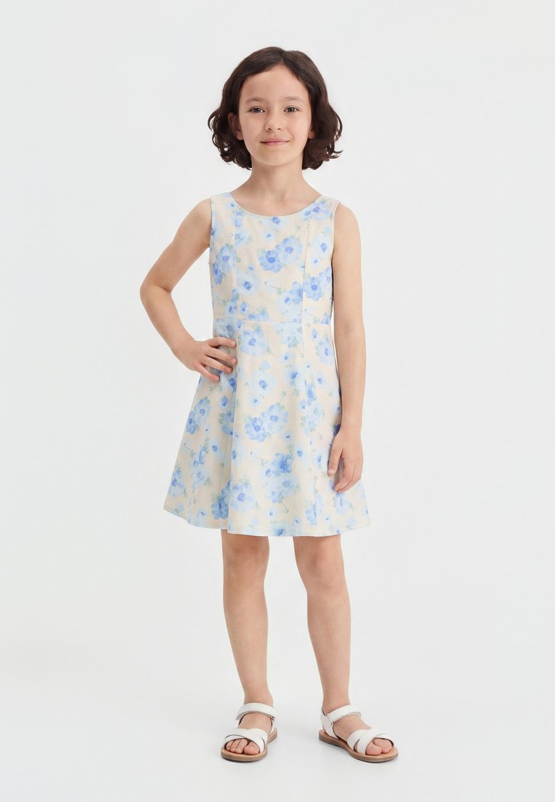 Fille aux cheveux courts et bruns portant une robe blanche sans manches avec des fleurs bleues et des sandales blanches, debout avec une main sur la hanche.