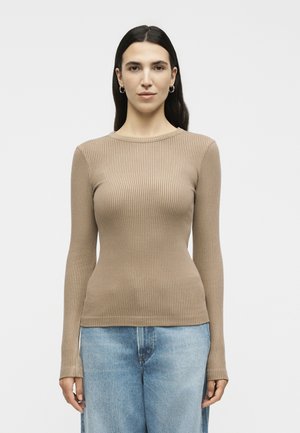 Γυναίκα με μακριά σκούρα μαλλιά, που φοράει μπεζ ριγωτό μακρυμάνικο τοπ και ανοιχτό μπλε τζιν, στέκεται μπροστά σε σκουρόχρωμο λευκό φόντο.