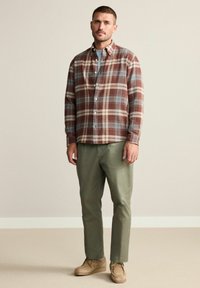 Camicia di flanella a quadri nei toni del marrone, crema e blu, con colletto button-down. Chinos verde oliva e scarpe beige completano l'outfit.