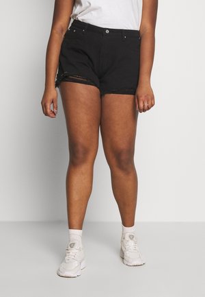 Persoon draagt zwarte gescheurde denimshorts, witte sneakers, witte sokken en een wit T-shirt, staand tegen een effen achtergrond.