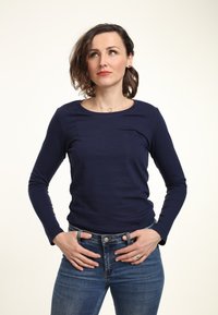 Granatowa koszula z długim rękawem z miękkiego, elastycznego materiału o okrągłym dekolcie. Noszona z niebieskimi dżinsami, ręce na biodrach, na jasnym tle.