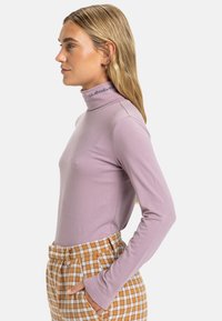 Maglione a maniche lunghe viola chiaro con collo a dolcevita in tessuto morbido, caratterizzato da un colletto a coste con logo del brand. Abbinato a pantaloni a quadri a vita alta.