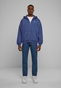 Hættezip-sweatshirt i mørkeblå, med frontlommer og broderet logo. Kombinationen er med lyseblå denim jeans og hvide sneakers.