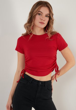 Červený rebrovaný crop top s krátkymi rukávmi, ktorý má bočné viazanie na prispôsobenie. Nosený s čiernymi džínami s vysokým pásom. Hladká textúra, priliehavý dizajn.