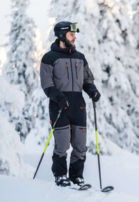 Man in black and gray ski suit holding poles, wearing helmet and goggles, standing on snow with snow-covered trees in the background.